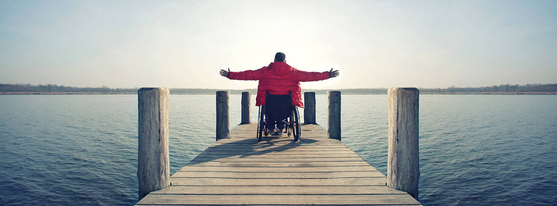 Hombre en silla de ruedas levantando los brazos en un muelle mirando al mar