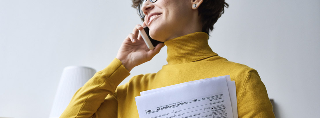 mujer con jersey amarillo hablando por teléfono con papeles de la declaración de la renta