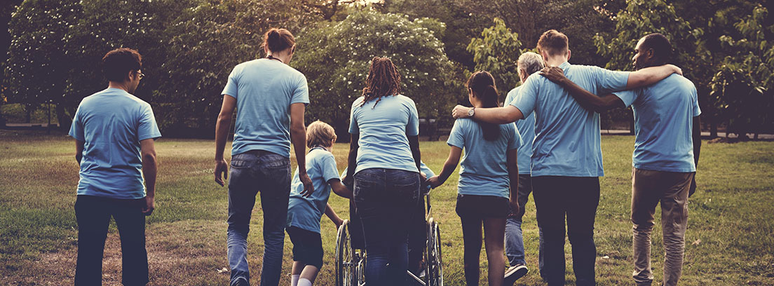 Grupo de voluntarios de distintas edades con camisetas azules pasean a una persona en silla de ruedas