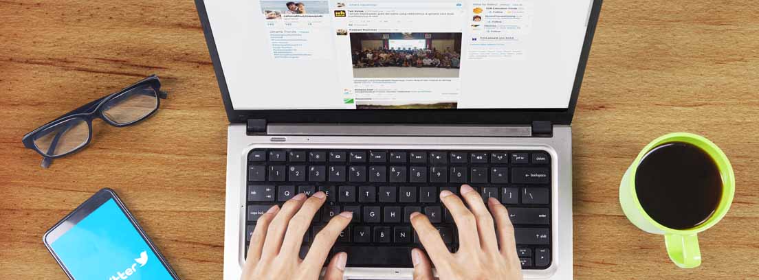 Vista cenital de unas manos sobre el teclado de un ordenador, una taza de café, unas gafas y un móvil con la pantalla de Twitter