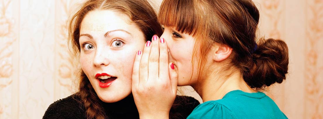 Una mujer contando un secreto al oído a otra mujer con gesto de sorpresa