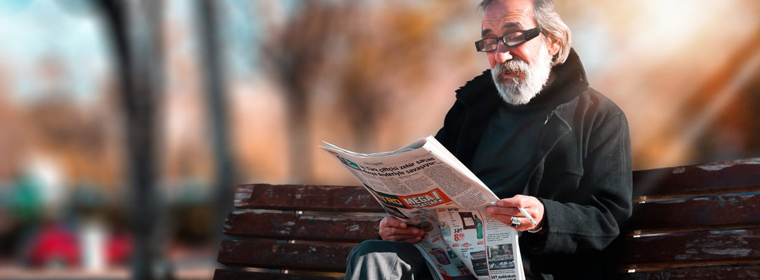 Hombre con barba y pelo blanco en banco de parque leyendo periódico