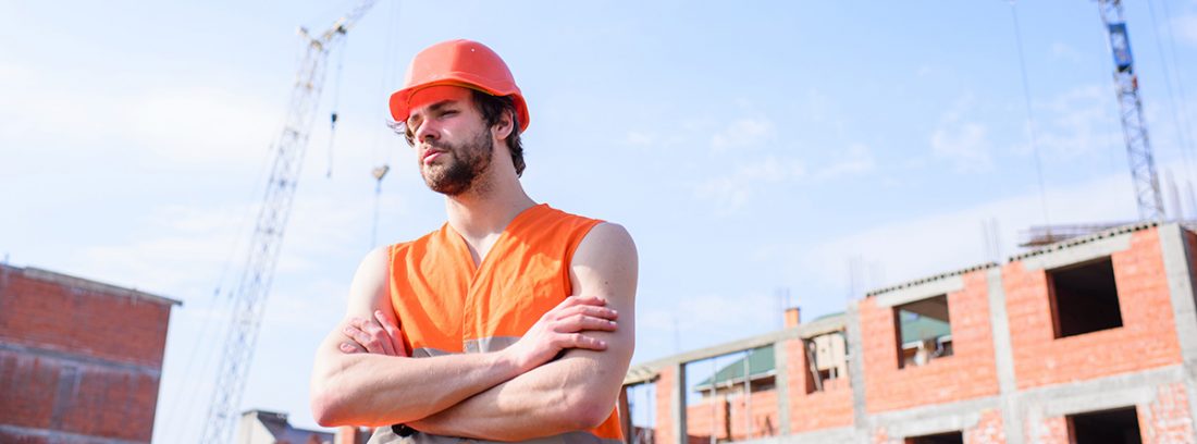 Hombre con casco rojo y chaleco naranja delante de unas viviendas en construcción